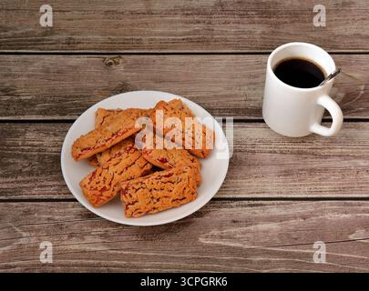 Piatto con una pila di biscotti fatti in casa freschi con marmellata di frutta su un tavolo di legno, accanto ad esso una tazza di caffè nero caldo. Primo piano. Foto Stock