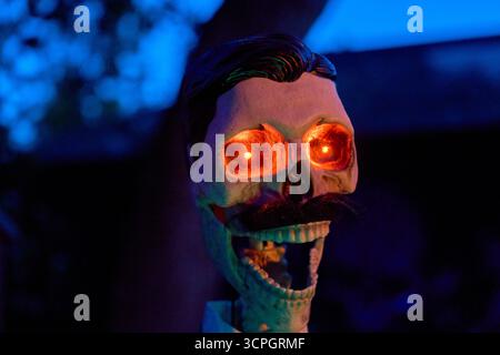 Ritratto ravvicinato di un cranio con baffi e occhi rossi accesi contro il crepuscolo blu. L'inquietante attrezzatura di Halloween crea un'atmosfera da cortile. Foto Stock
