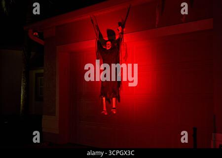Un mietitore con cappuccio e gambe ossee è appeso alla porta di un garage sotto una luce rossa profonda. Spettrale scenografia da cortile di Halloween con ombre spettacolari di notte. Foto Stock