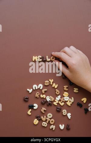 Primo piano della mano che organizza un saluto da lettere di plastica sparse su sfondo marrone. Concetto creativo, struttura piatta, spazio di copia Foto Stock