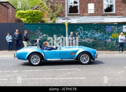 Uomo che guida una replica di auto sportiva AC Cobra blu d'epoca su Morpeth Street con spettatori e pareti coperte di graffiti sullo sfondo Foto Stock