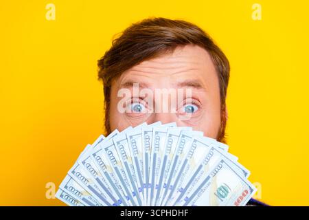 Un giovane eccitato che tiene in mano un fan del denaro contante su un vivace sfondo giallo, mostrando sorpresa e successo Foto Stock