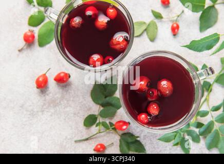 Tè caldo alle cime di rosa in tazze di vetro con rosa fianchi freschi e foglie verdi su sfondo bianco Foto Stock