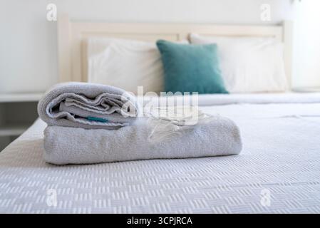 La camera da letto è dotata di un letto fatto con fresche lenzuola bianche. Un asciugamano ben arrotolato siede sul letto, accompagnato da un tocco di blu da un de Foto Stock