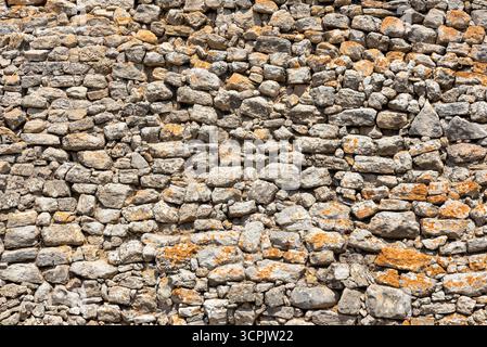 Varie pietre formano una parete ruvida e ruvida, che mostra sfumature di grigio e marrone. Il vivace lichene arancione contrasta splendidamente con il paesaggio intramontabile Foto Stock