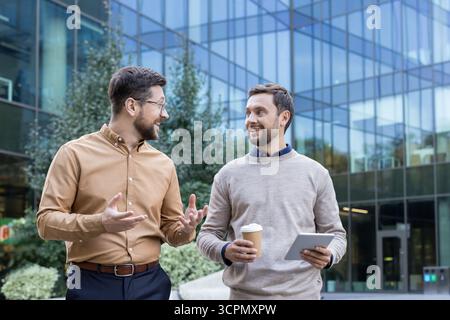 Due allegri uomini d'affari che camminano e discutono attivamente di un progetto o di nuove idee, portando un tablet e una tazza di caffè fuori da un moderno edificio di vetro Foto Stock