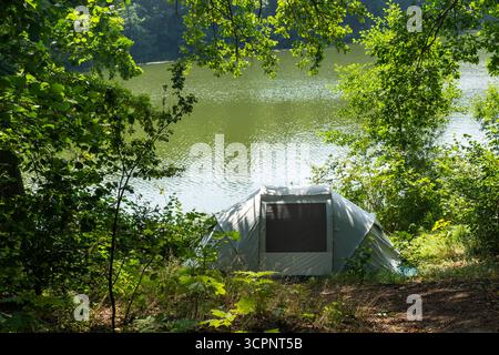 Tenda da da campeggio grigia allestita da un tranquillo lago circondato da lussureggianti alberi di foresta il giorno d'estate. Concetto di campeggio nella natura selvaggia, avventura all'aperto e Foto Stock
