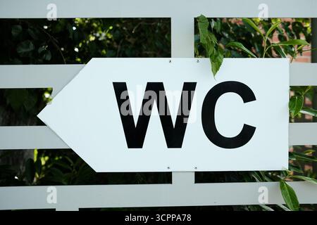 Cartello direzionale bianco con lettere WC nere rivolte a sinistra, montato su una recinzione con foglie verdi sullo sfondo. Indicazione toilette esterna in Foto Stock