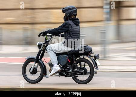 Giovane motociclista in abbigliamento casual e casco cavalca la moto nera lungo la strada urbana. Lo scatto dinamico con sfocatura in movimento enfatizza velocità, energia e. Foto Stock