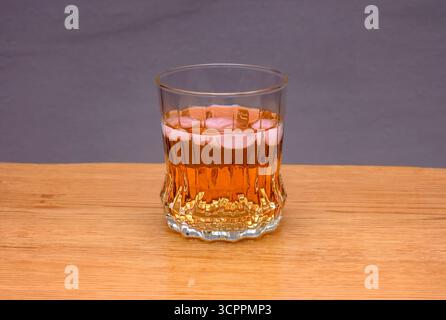 Un vetro trasparente riempito di liquido color ambra e cubetti di ghiaccio è posizionato su un tavolo di legno chiaro su uno sfondo grigio neutro. Foto Stock