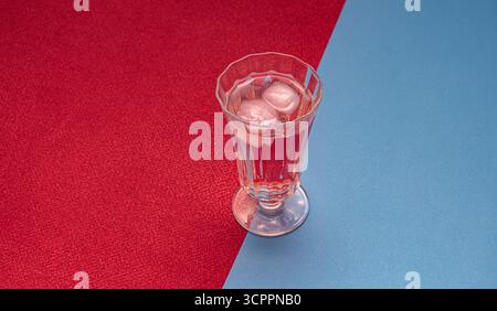 Un elegante bicchiere riempito con una bevanda rosata e cubetti di ghiaccio è collocato su uno sfondo rosso e blu diagonalmente diviso. Il contrasto tra il freddo. Foto Stock