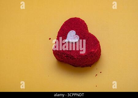 Torta in velluto rosso a forma di cuore su una superficie ruvida. Piccolo cuore bianco decorativo sulla parte superiore. San Valentino, romanticismo e dolce indulgenza. Foto Stock