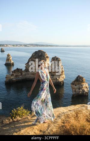 Bella donna con un lungo abito floreale estivo che si trasforma e si gode di splendide formazioni rocciose e il tramonto sul mare a Lagos in Portogallo. Modello alla moda Foto Stock