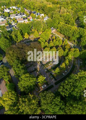 Vista aerea di filari curvi di cimiteri circondati da una fitta foresta, con cappella al centro e case residenziali vicine, che mescolano natura e design umano. Foto Stock