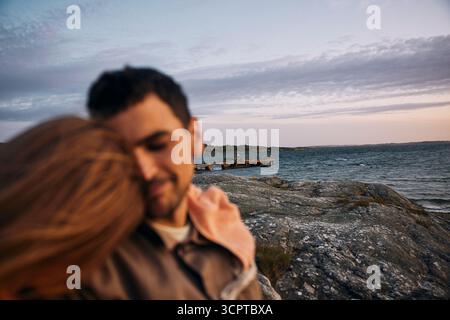 Coppia affettuosa defocalizzata con una tranquilla scena di mare vicino alle rocce sullo sfondo al tramonto Foto Stock