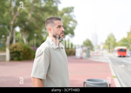 Un uomo con la testa rasata e la barba sta su un marciapiede accanto a un bidone. La scena è ambientata in una città, con un autobus sullo sfondo. Quell'uomo è wa Foto Stock