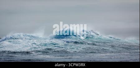 Una grande onda si schianta nell'oceano, la sua acqua turchese spruzza nell'aria contro un cielo grigio, mostrando la potenza grezza e la bellezza della natura. Foto Stock