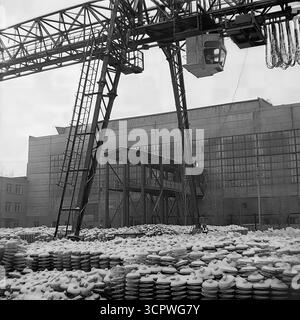 Una gru a cavalletto con operatore in cabina si trova su un cortile pieno di isolanti in ceramica ricoperti di neve presso lo Slavyansk Fittings and Insulator Plant (AIZ) nella RSS Ucraina negli anni '1970 Questa suggestiva foto d'archivio monocromatica cattura una scena invernale in una delle principali imprese industriali sovietiche. Il vasto inventario di prodotti finiti, pronti per la spedizione nonostante la neve, mostra le dimensioni della produzione di massa nell'industria pesante della regione del Donbas. Foto Stock