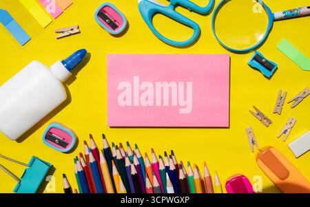 Composizione verticale di vari articoli di cancelleria scolastica che circondano un foglio di carta rettangolare rosa brillante su uno sfondo giallo brillante. Perfetto per promozioni back-to-School, design a tema scolastico o banner didattici con spazio di testo per il testo. Foto Stock
