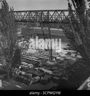 Il deposito all'aperto dello Slavyansk Fittings and Insulator Plant (AIZ) è pieno di migliaia di isolanti in ceramica finiti nella RSS Ucraina negli anni '1970 Questa foto d'archivio monocromatica cattura l'immensa scala della produzione di massa dell'era sovietica, con prodotti impilati ordinatamente in casse e su pallet. Una grande gru a cavalletto attraversa il cantiere, utilizzata per la logistica e la spedizione di questi componenti essenziali per i progetti di elettrificazione dell'URSS, mostrando la potenza industriale della regione del Donbas. Foto Stock