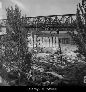 Il deposito all'aperto dello Slavyansk Fittings and Insulator Plant (AIZ) è pieno di migliaia di isolanti in ceramica finiti nella RSS Ucraina negli anni '1970 Questa foto d'archivio monocromatica cattura l'immensa scala della produzione di massa dell'era sovietica, con prodotti impilati ordinatamente in casse e su pallet. Una grande gru a cavalletto attraversa il cantiere, utilizzata per la logistica e la spedizione di questi componenti essenziali per i progetti di elettrificazione dell'URSS, mostrando la potenza industriale della regione del Donbas. Foto Stock