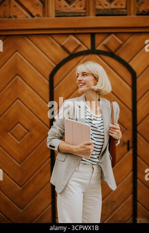 Una donna sicura di sé si trova davanti a una porta di legno, con un blazer e una camicia a righe. Tiene in mano un tablet e una borsa, sorridendo brillantemente in una giornata calda, fa la mostra Foto Stock