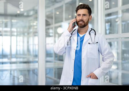 Medico maschile in camice bianco e stetoscopio che comunica sul suo smartphone, che rappresenta la comunicazione sanitaria, la telemedicina e la consulenza medica professionale in un ambiente clinico Foto Stock