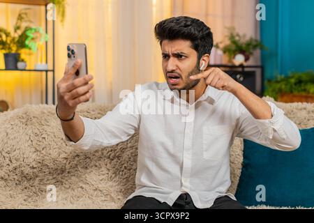 Un uomo indiano a casa sul divano che discute durante una videochiamata, parlando attraverso le cuffie. Ragazzo arabo frustrato, emotivo, alzare la voce, lo stress, reagire intensamente, esprimere disaccordo e tensione Foto Stock