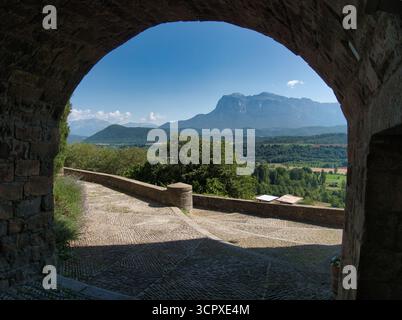 Vista della Valle delle montagne attraverso l'antico Arco di pietra - Una vastissima valle illuminata dal sole e maestose montagne blu incorniciate da un arco di pietra rustico scuro e acciottolato Foto Stock