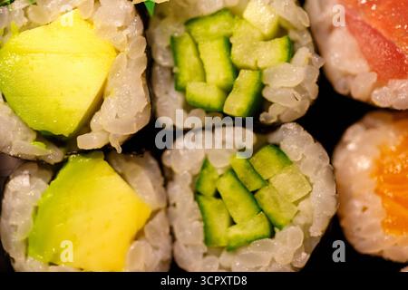 Vista ravvicinata dall'alto di vari sushi roll. I panini presentano riso bianco, cetriolo verde, avocado giallo e pesce crudo di arancia rosa. Foto Stock