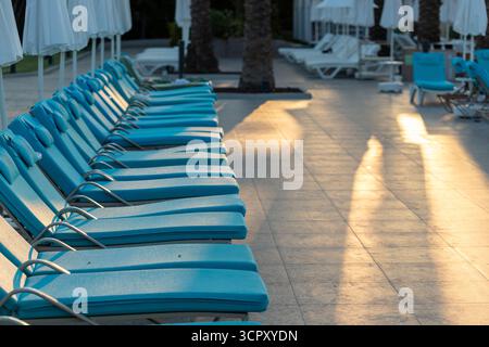 Fila di lettini blu con ombrelloni bianchi vicino alle palme su un terreno piastrellato con lunghe ombre dalla luce del sole. Foto Stock