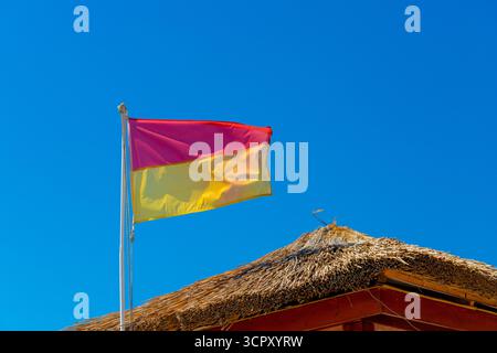 Una bandiera bagnino, rossa su gialla, ondeggia contro un cielo azzurro. Edificio con tetto in paglia visibile. Foto Stock