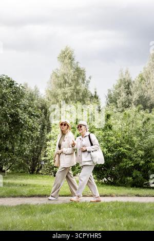 Due donne anziane sicure che camminano all'aperto a tutta lunghezza, abbigliamento casual elegante con cappelli e borse, che rappresentano uno stile di vita positivo all'età, auto conf Foto Stock