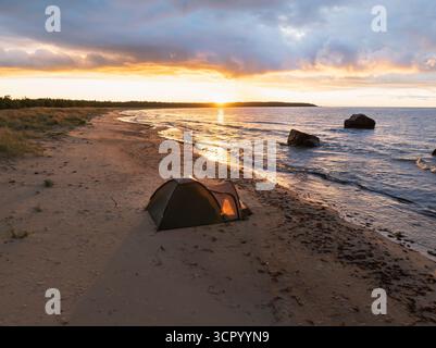 Campeggio tranquillo su una spiaggia di sabbia selvaggia con una tenda illuminata che si affaccia sul mare durante un tramonto dorato mozzafiato. Foto Stock
