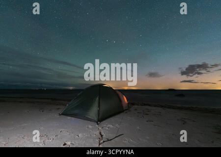 Un'accogliente tenda da campeggio illuminata poggia su una spiaggia sabbiosa sotto un cielo notturno mozzafiato e pieno di stelle sul mare. Foto Stock