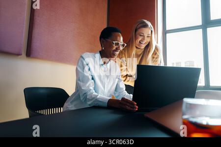 Due donne in un ufficio moderno sorridono mentre sono impegnate nel lavoro utilizzando un notebook. Sembrano concentrati, di sostegno e amano collaborare su un progetto che io Foto Stock