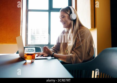 Una giovane donna con lunghi capelli biondi è seduta al chiuso e utilizza il suo computer portatile indossando le cuffie. Sorride e partecipa a una riunione online o c Foto Stock