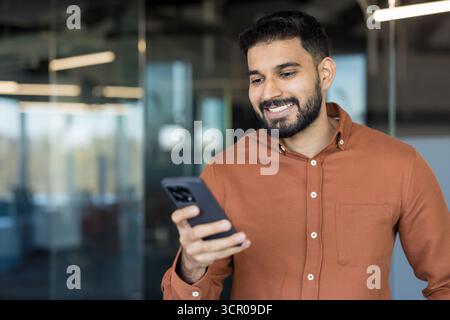 Giovane uomo d'affari indiano sorridente in generale mentre tiene e guarda uno smartphone, si occupa di comunicazione digitale, lavoro online o social media in un ambiente di ufficio moderno Foto Stock