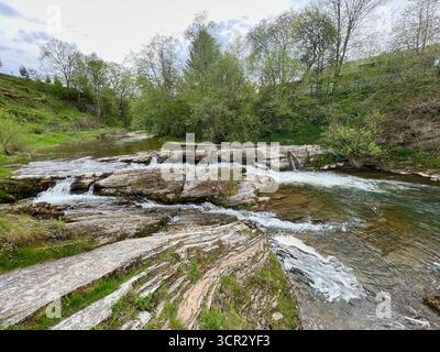 Il pittoresco ruscello scorre su rocce piane e stratificate, formando dolci cascate in un paesaggio lussureggiante e verde. Alberi e arbusti che aggiungono bellezza naturale alla scena. L'acqua limpida riflette l'ambiente circostante sereno. Foto Stock