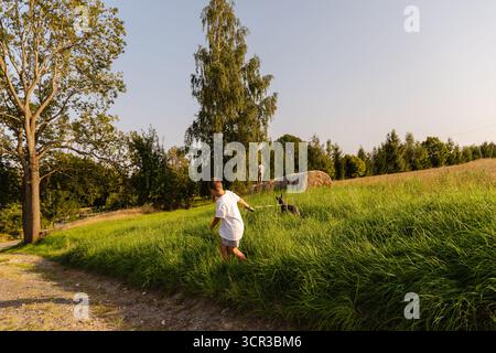 Un uomo cammina accanto a suo figlio mentre passeggia attraverso un lussureggiante campo verde. Il loro cane corre giocosamente avanti. Il sole splende brillantemente, emettendo un caldo bagliore Foto Stock