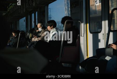 I passeggeri si siedono in una tranquilla contemplazione su un treno di Osaka, i loro volti sono parzialmente illuminati da forti raggi di luce solare Foto Stock