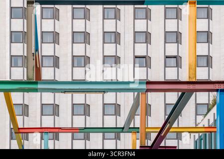 Struttura colorata in metallo astratto con travi e travi intersecanti poste contro la ripetitiva facciata in pietra di un moderno edificio per uffici Foto Stock