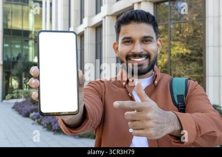 Uomo indiano che presenta uno schermo vuoto per smartphone con un gesto di puntamento, pronto per contenuti personalizzati mentre è in piedi all'aperto, che rappresenta la tecnologia moderna e i concetti di comunicazione Foto Stock