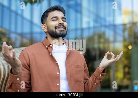 Uomo indiano che medita all'aperto in stile zen, tiene gli occhi chiusi, si concentra sul respiro e pratica la consapevolezza per l'equilibrio mentale e il benessere emotivo Foto Stock