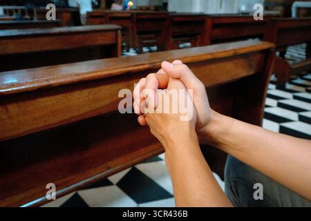 visto da un lato un uomo tiene le dita intrecciate in preghiera all'interno della chiesa circondato da panche e dal pavimento bianco e nero a motivi geometrici Foto Stock