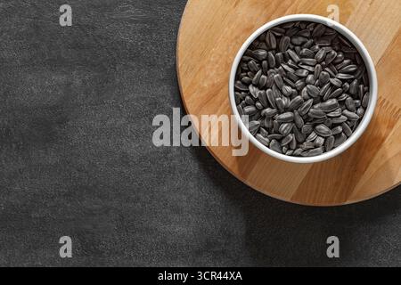 semi di girasole neri non sbucciati nel recipiente sul piano di lavoro scuro, vista dall'alto Foto Stock