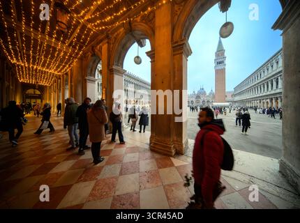 I visitatori passeggiano attraverso i portici di Piazza San Marco, mentre le luci festose brillano sopra e il Campanile si erge alto sullo sfondo. Foto Stock