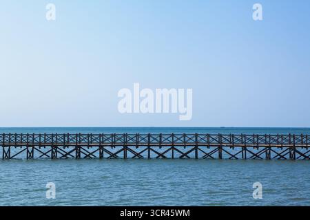 Lungo ponte in legno si estende attraverso le calme acque turchesi. Tradizionale design indonesiano, tranquillo paesaggio marino, cielo azzurro e limpido creano un'atmosfera tropicale Foto Stock