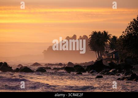 Tramonto vibrante su una spiaggia rocciosa con palme sagomate e nebbia lontana. Krui, Lampung, Sumatra, Indonesia Foto Stock