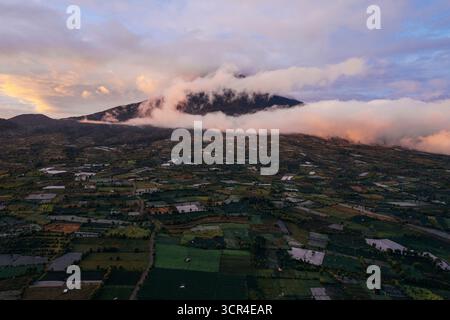 Vista aerea di lussureggianti campi verdi con un vulcano avvolto da nuvole al tramonto. Kayu Aro, Kerinci Regency, Jambi, Sumatra, Indonesia Foto Stock